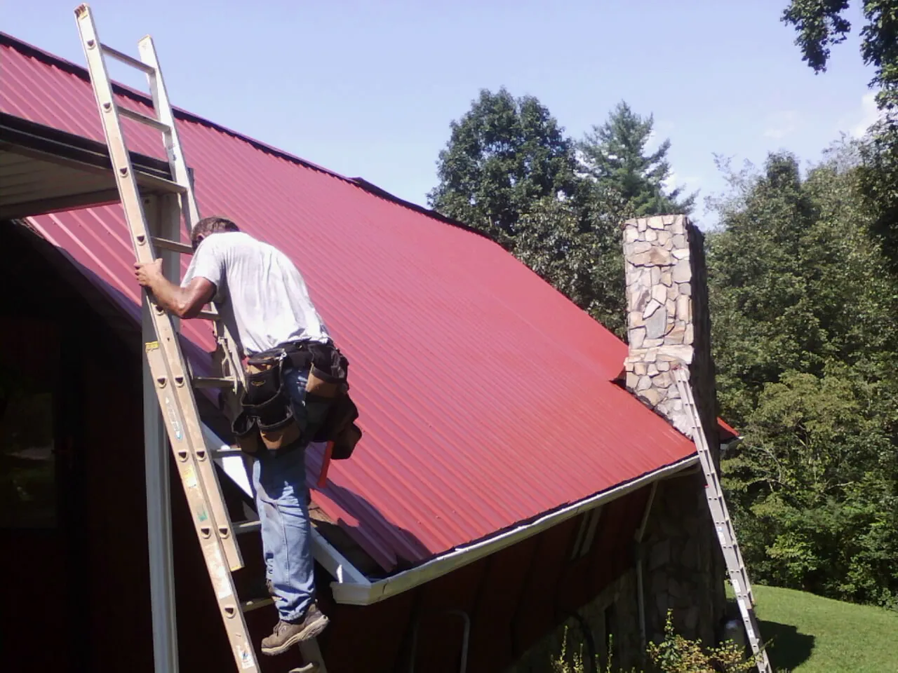 Roofer on a ladder beside a red metal roof and stone chimney — Soffit & Fascia Repair by Roofing Brothers in New Wilmington